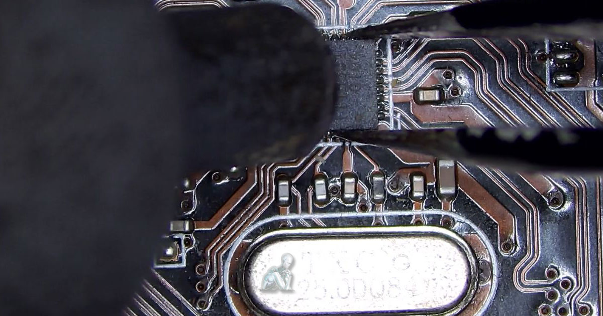 Technician using hot air to remove a central IC chip from a motherboard while holding it with tweezers, with a crystal oscillator visible beneath during component-level repair