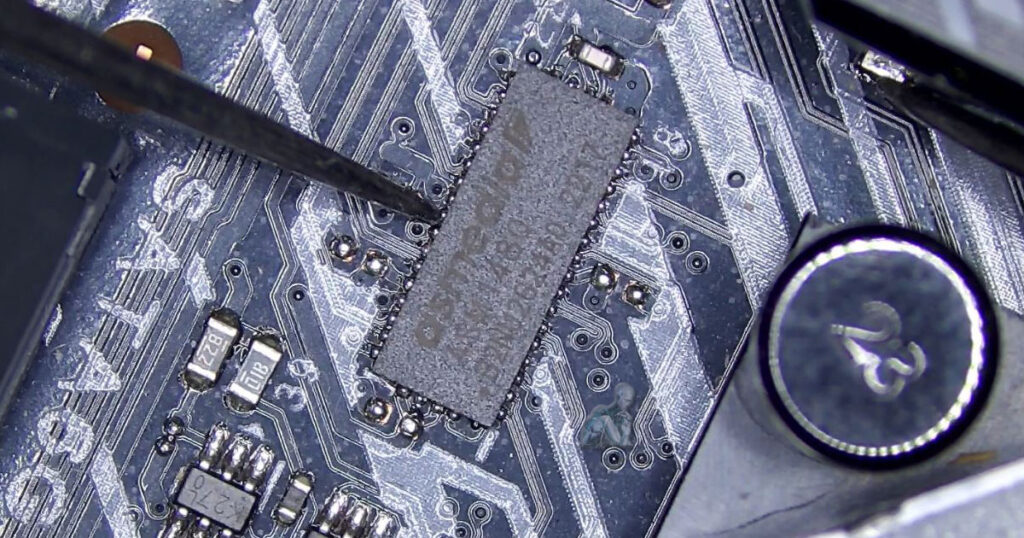 Board-level view of a DS media IC on a motherboard while multimeter probes contact its pins to test for a short, with surrounding capacitors and signal traces visible