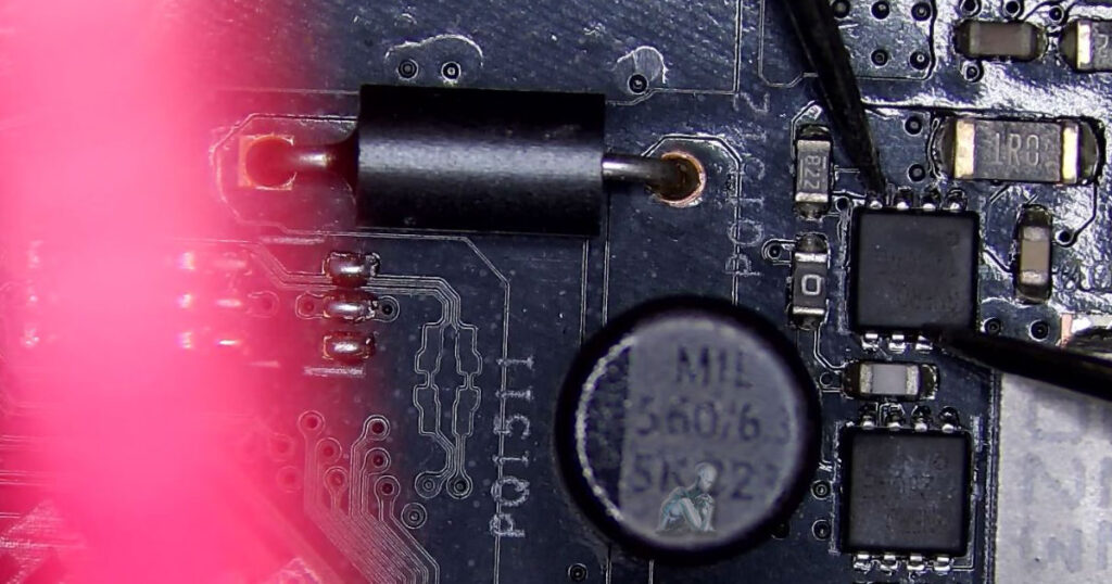 Inspection scene showing multimeter probes placed on MOSFET terminals to test for a short, with nearby capacitors, ICs, and circuit traces visible