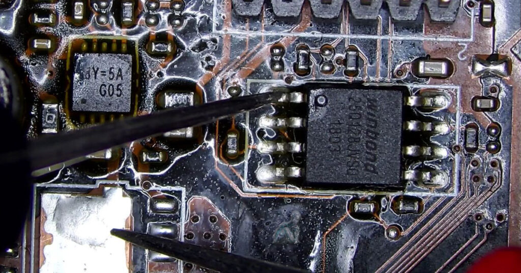 Technician probing a leg of a BIOS chip to ground on a motherboard to check for a short circuit during diagnostic testing