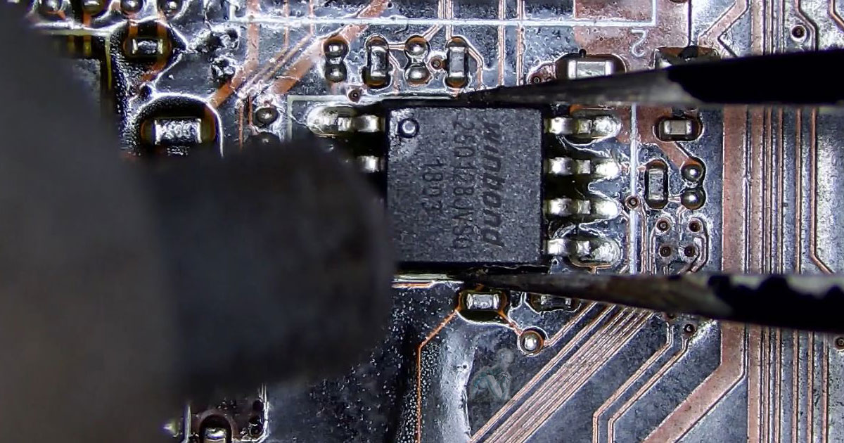 Hot air tool and tweezers lifting a BIOS chip from a motherboard during removal, with surrounding components exposed on the circuit board