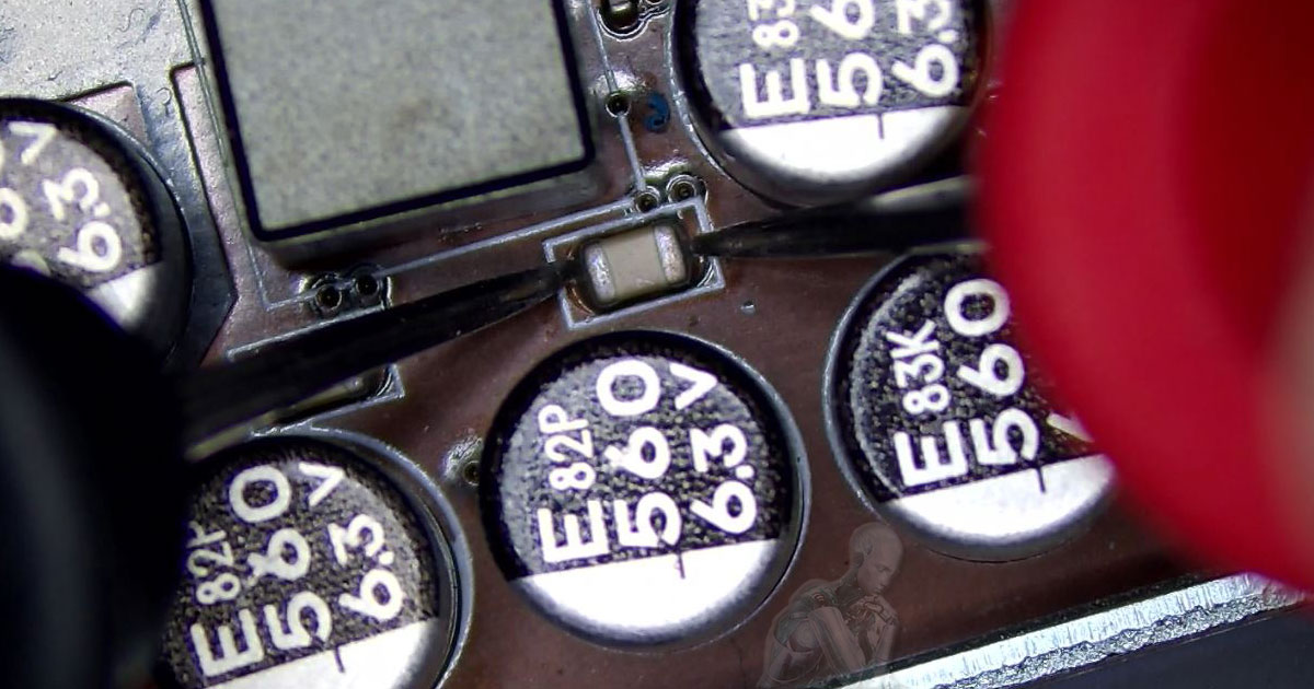 Probe testing a small surface mount capacitor on a motherboard, partially hidden between four larger 560µF 6.3V electrolytic capacitors clustered around it.