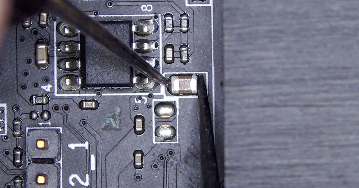 Measurement scene with multimeter probes placed across a capacitor to confirm a short, with adjacent resistors, ICs, and solder connections visible