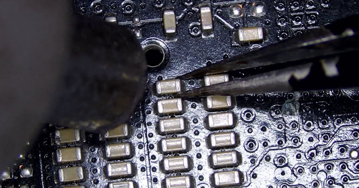 Hot air rework process heating an SMD capacitor on a circuit board as it is lifted from its pads, with nearby microcomponents and solder points visible
